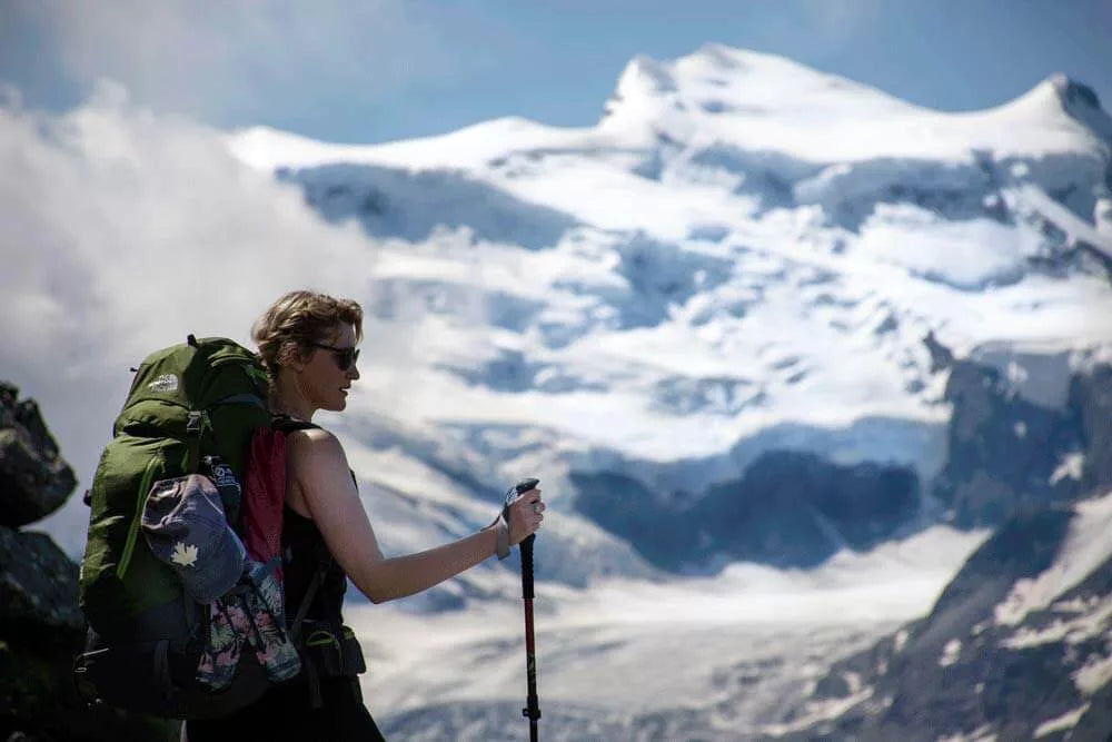 Person hiking with a backpack and walking stick in front of a snow-capped mountain.