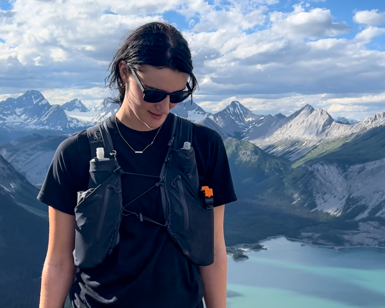 A lady wearing sunglasses standing in front of a mountainous landscape with a lake